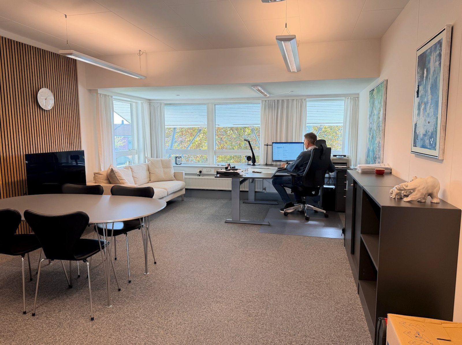 Kontorlandskab med mødebord, hvid sofa og arbejdsplads ved vindue